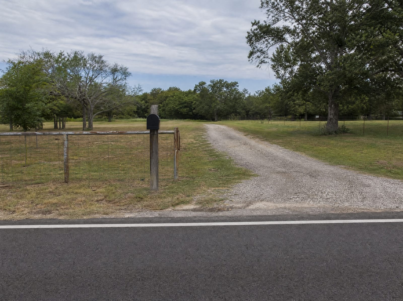 A Texas property with road frontage along a two-lane road, a gravel driveway and a wire fence line that a taking could affect, open land under flat daylight