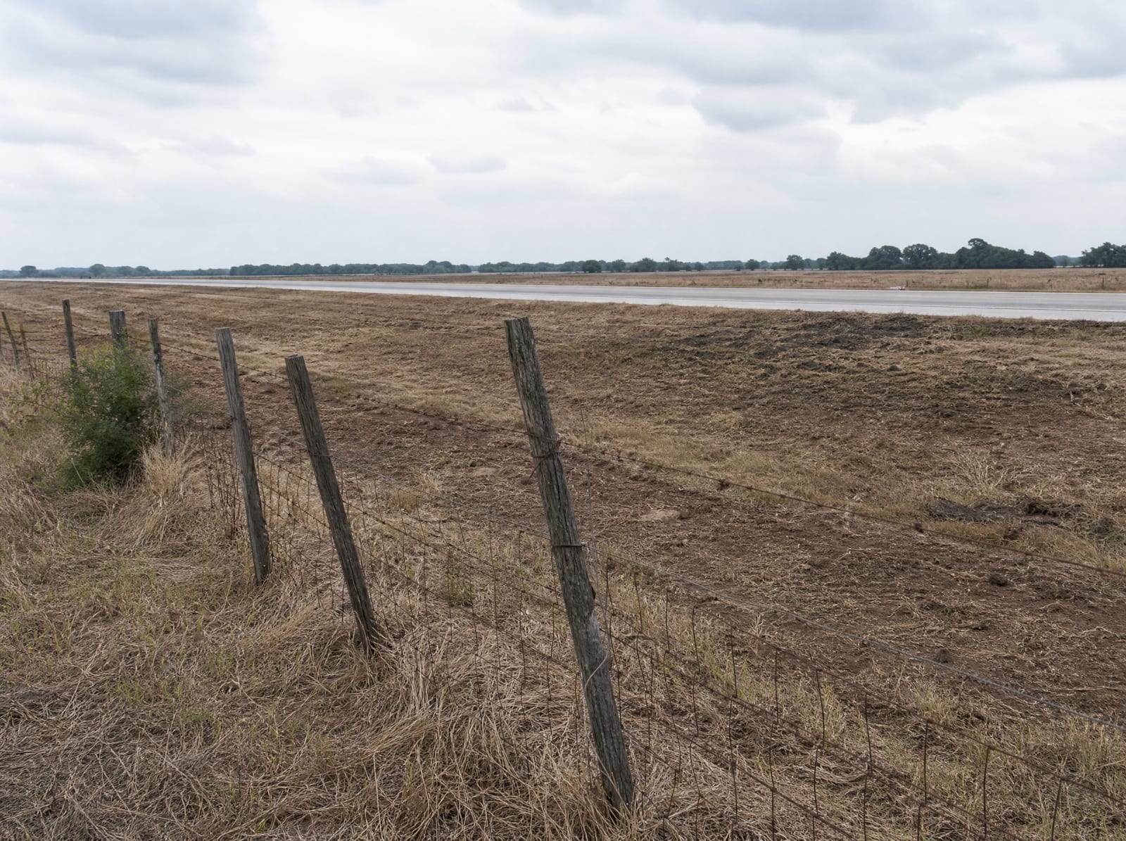 A Texas property and weathered wire fence line at the edge of a public project corridor, open land and a graded right-of-way in the distance under flat daylight