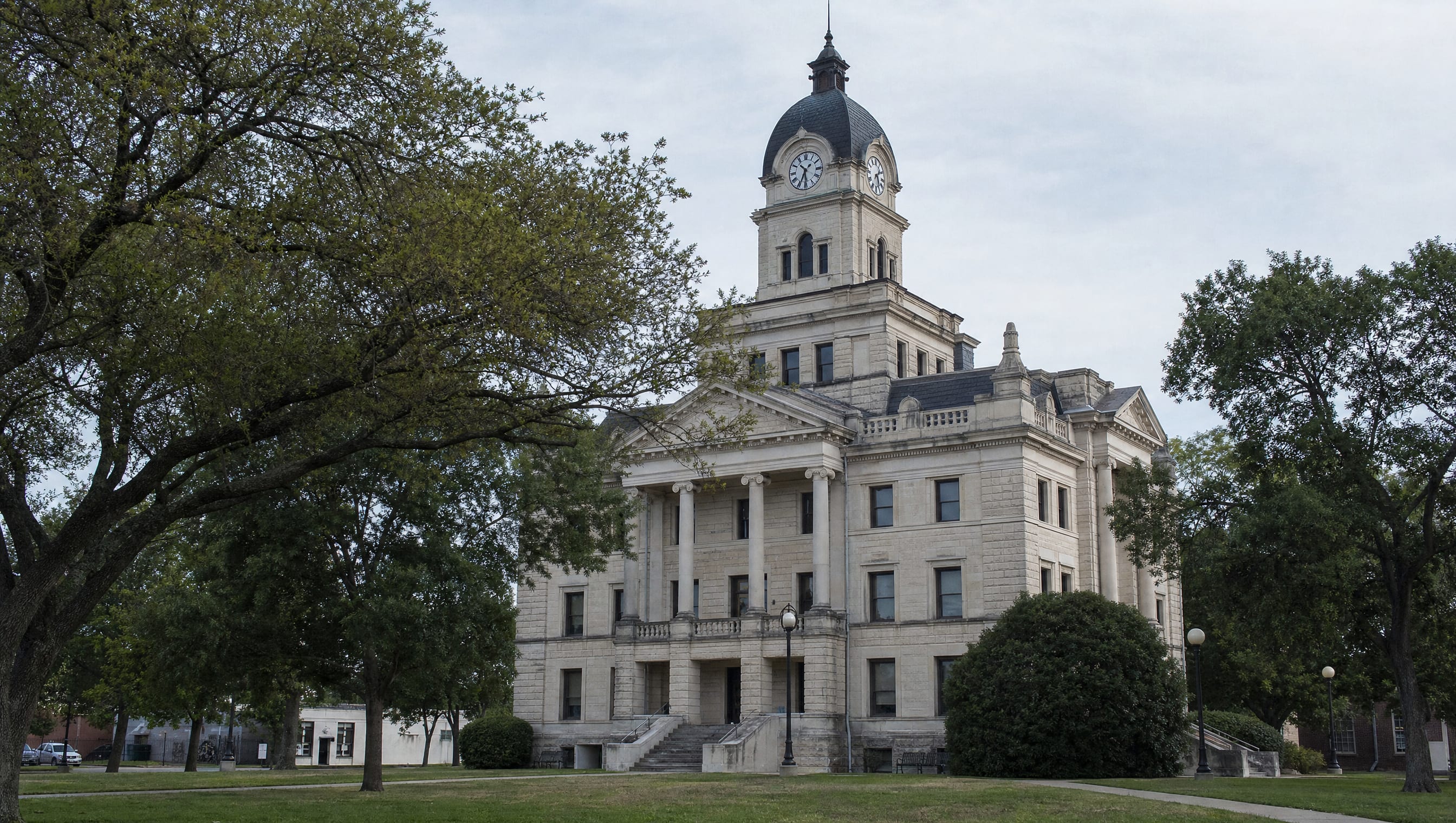 A stately Texas county courthouse of limestone with a clock tower, columns, and stone steps among oak trees on the courthouse lawn