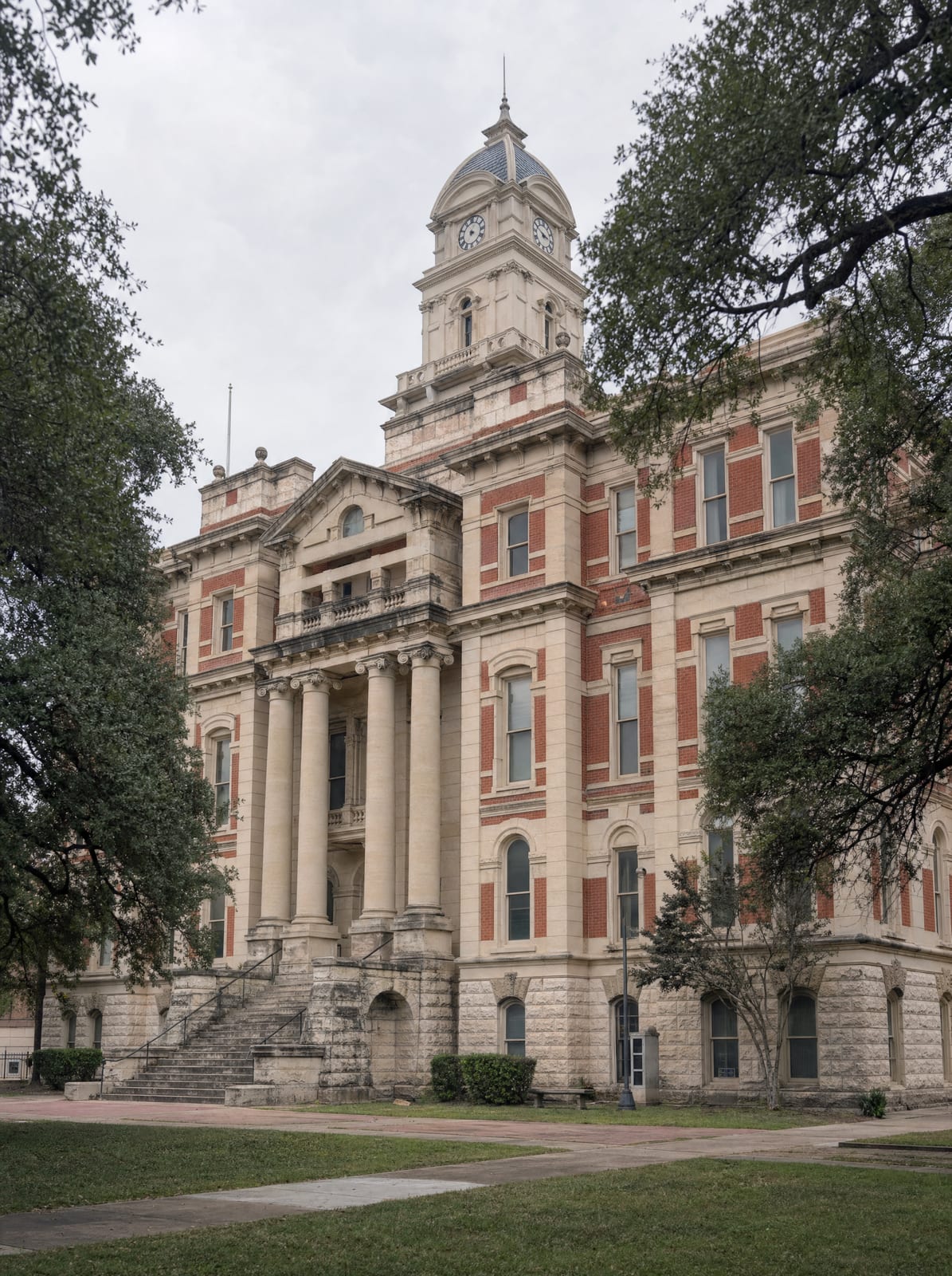 A dignified historic Texas county courthouse of warm limestone and red brick with a clock tower, columns, and stone steps among live oaks, for commercial and contract litigation