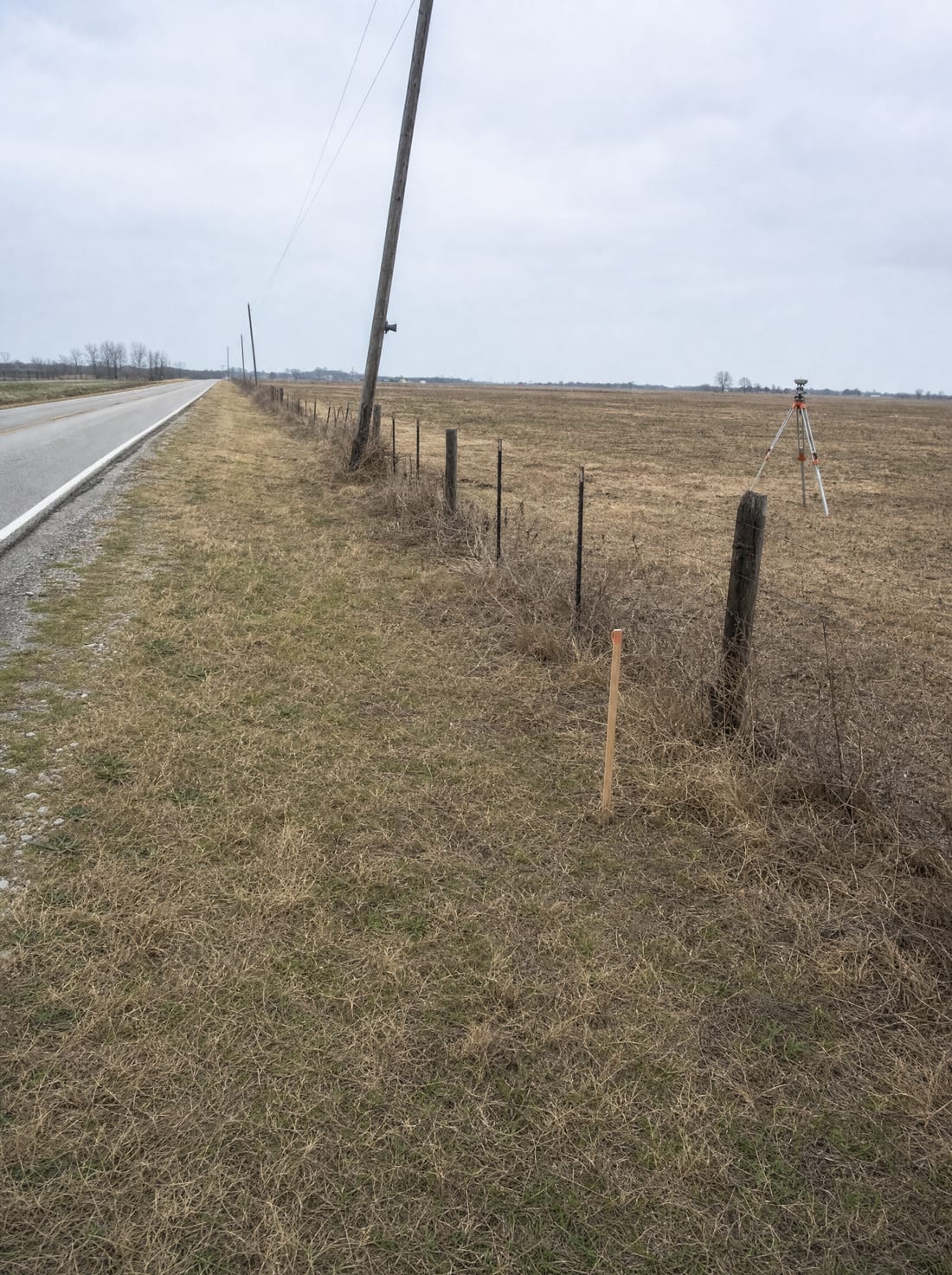 A flat rural Texas parcel along a barbed-wire fence with a land surveyor's tripod and stake in the field, for the initial notice and survey