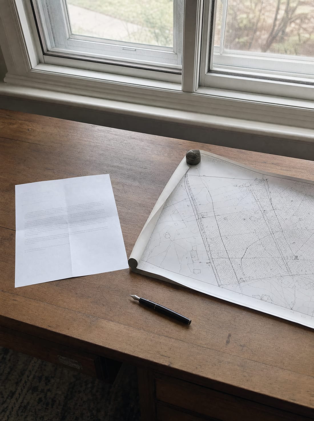 A desk by a window with an offer letter beside a rolled-out property parcel map and a pen, for the offer and negotiation period