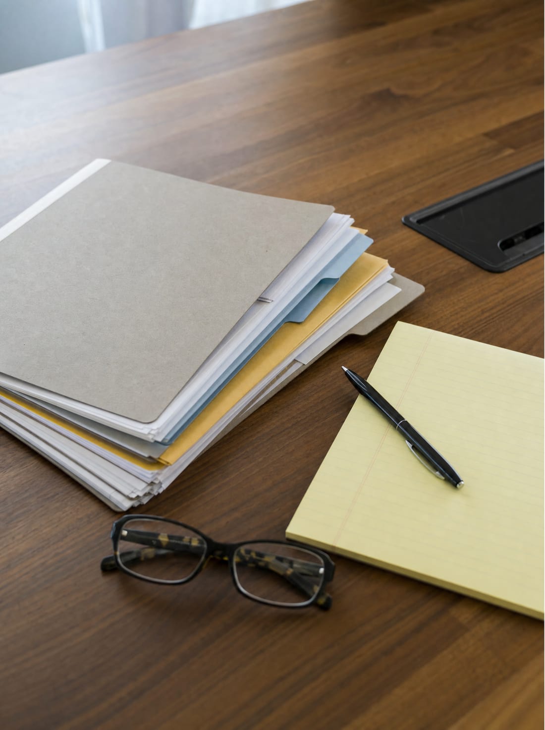 An organized folder of documents, a notepad, a pen, and reading glasses laid out on a wood desk