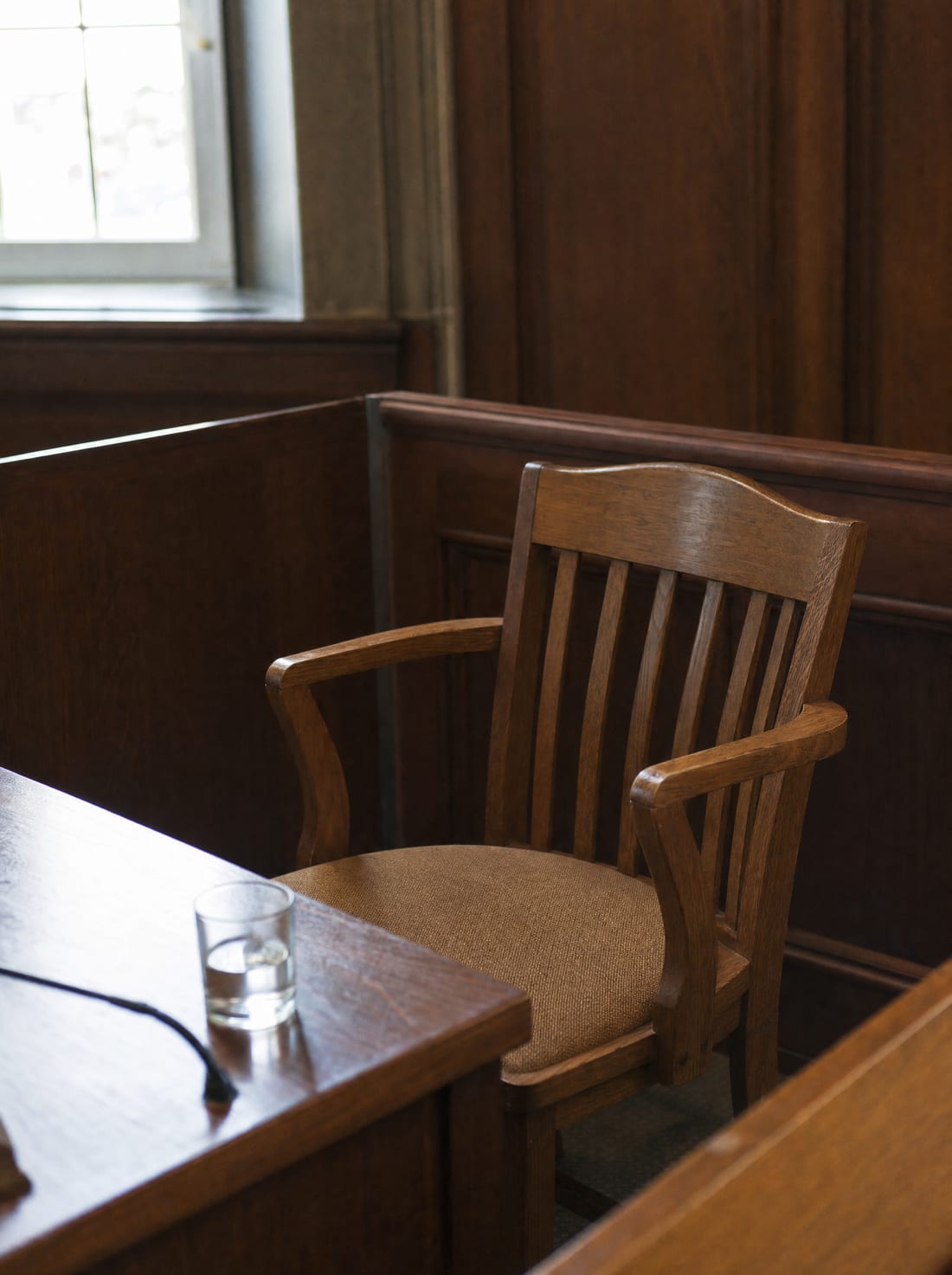 A single empty wooden witness chair beside a small table in a wood-paneled hearing room