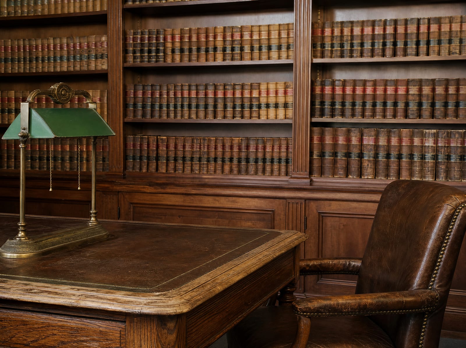A distinguished law-firm library of well-worn bound law reporters with a wood reading table, a green-shaded brass banker's lamp, and a leather chair