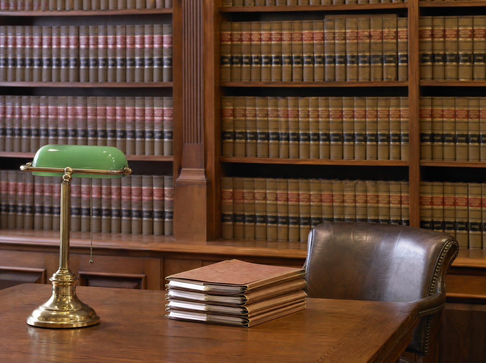 A distinguished, established law-firm library of well-worn bound law reporters with a wood reading table, a green-shaded brass banker's lamp, a leather chair, and closed case files, conveying experience and trust