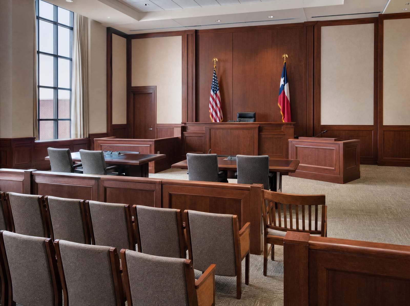 An orderly courtroom with a judge's bench, a witness stand, wood-paneled walls, a United States flag and a Texas flag, and rows of seating, where a commercial case goes to trial