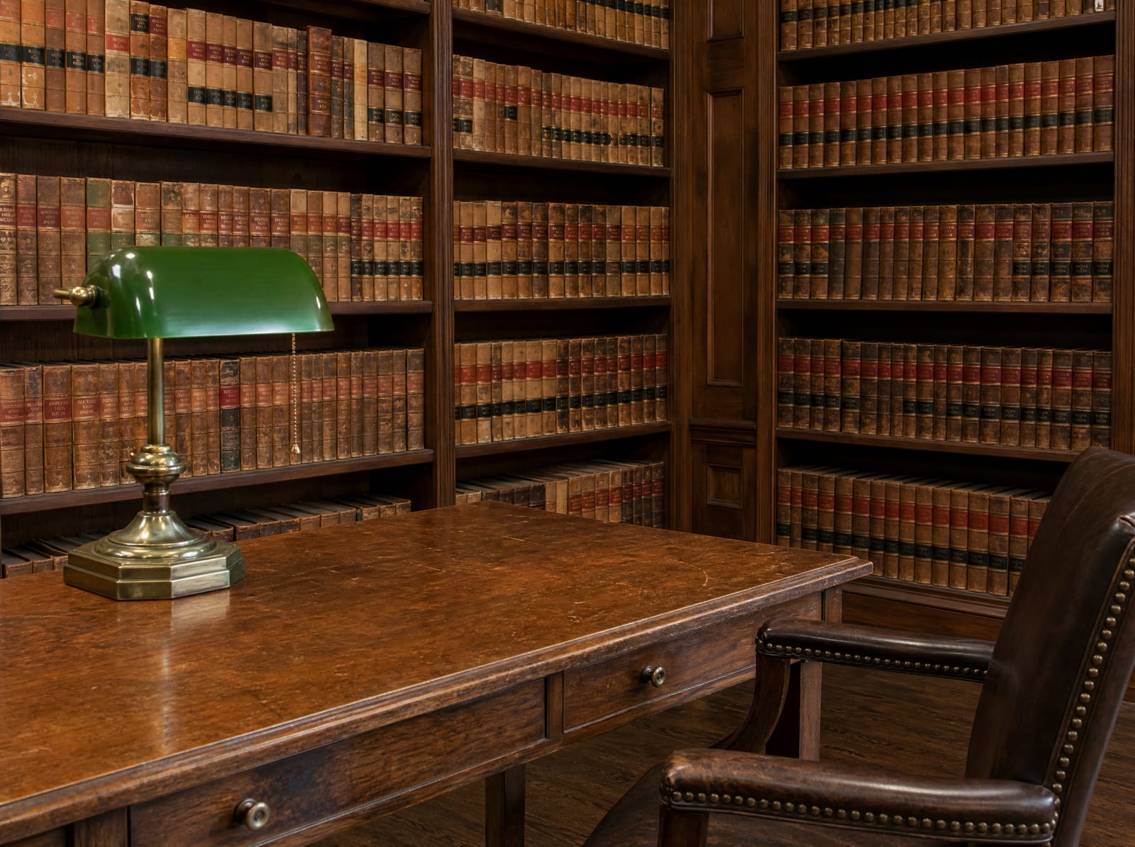 A distinguished law-firm library of well-worn bound law reporters with a wood reading table, a green-shaded brass banker's lamp, and a leather chair
