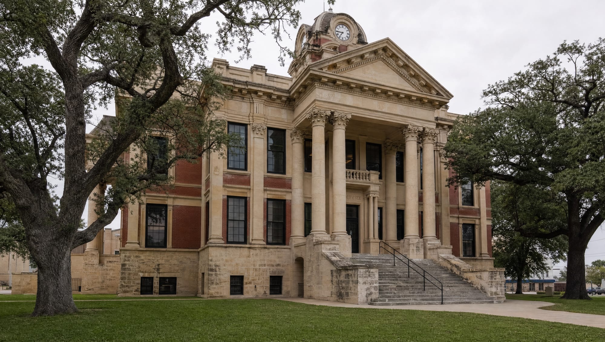 A dignified historic Texas county courthouse of warm limestone and red brick with a clock tower, columns, and broad stone steps among live oaks, where commercial and contract disputes are decided