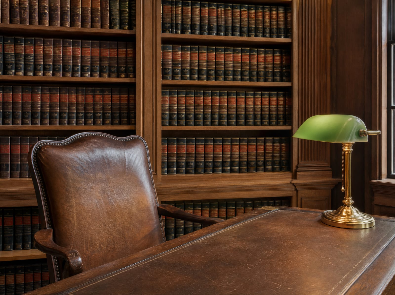 A distinguished law-firm library of well-worn bound law reporters with a wood reading table, a green-shaded brass banker's lamp, and a leather chair