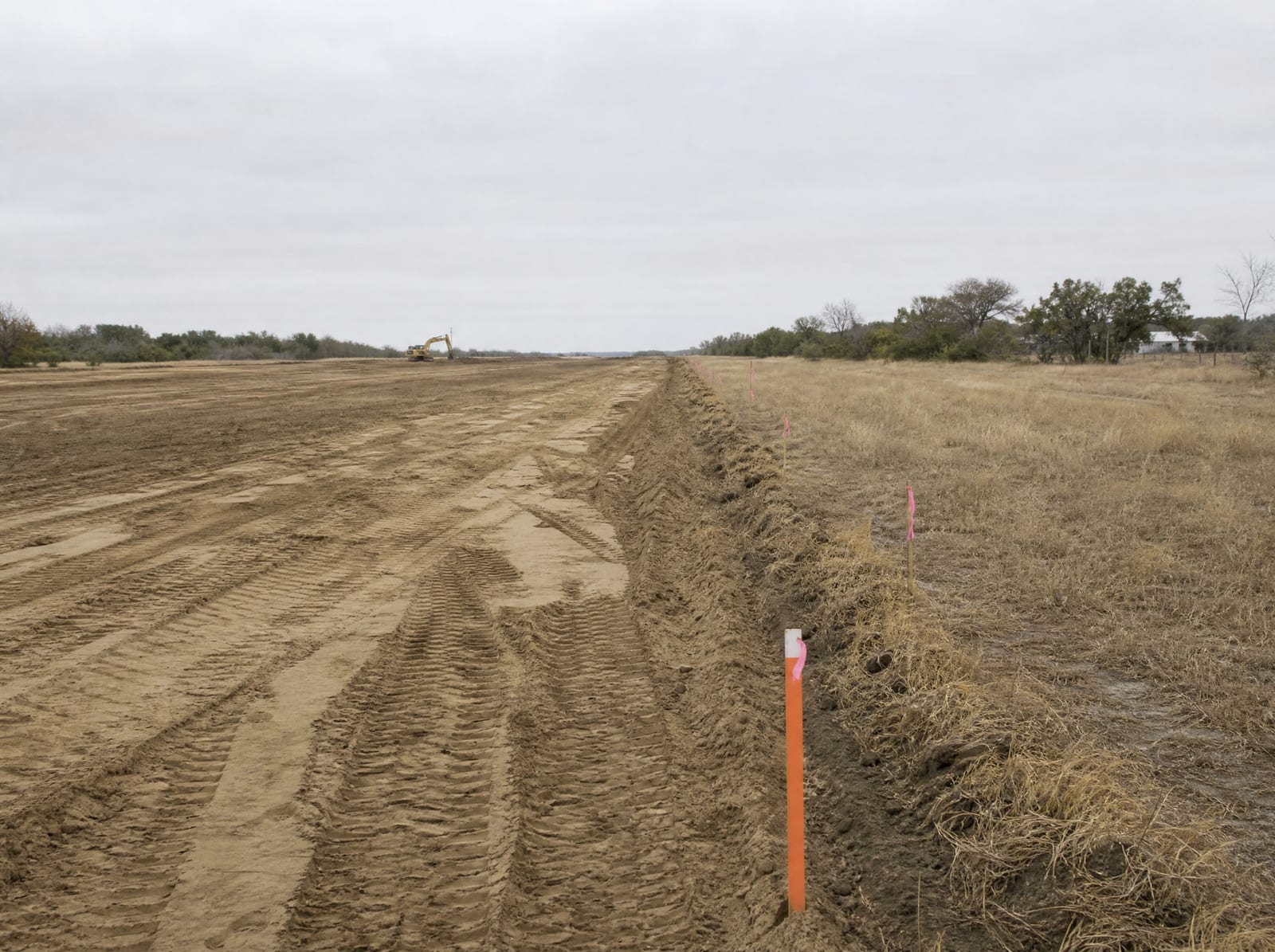 An active infrastructure construction site of graded earth and a partial roadbed across open land, with survey stakes and right-of-way flags along the property boundary and a distant excavator