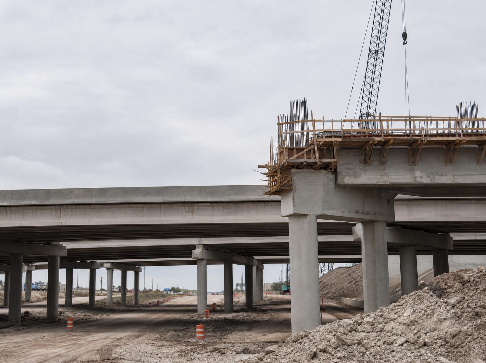 A concrete highway overpass or interchange under construction with support columns, exposed rebar, formwork, and a crane over a graded roadway