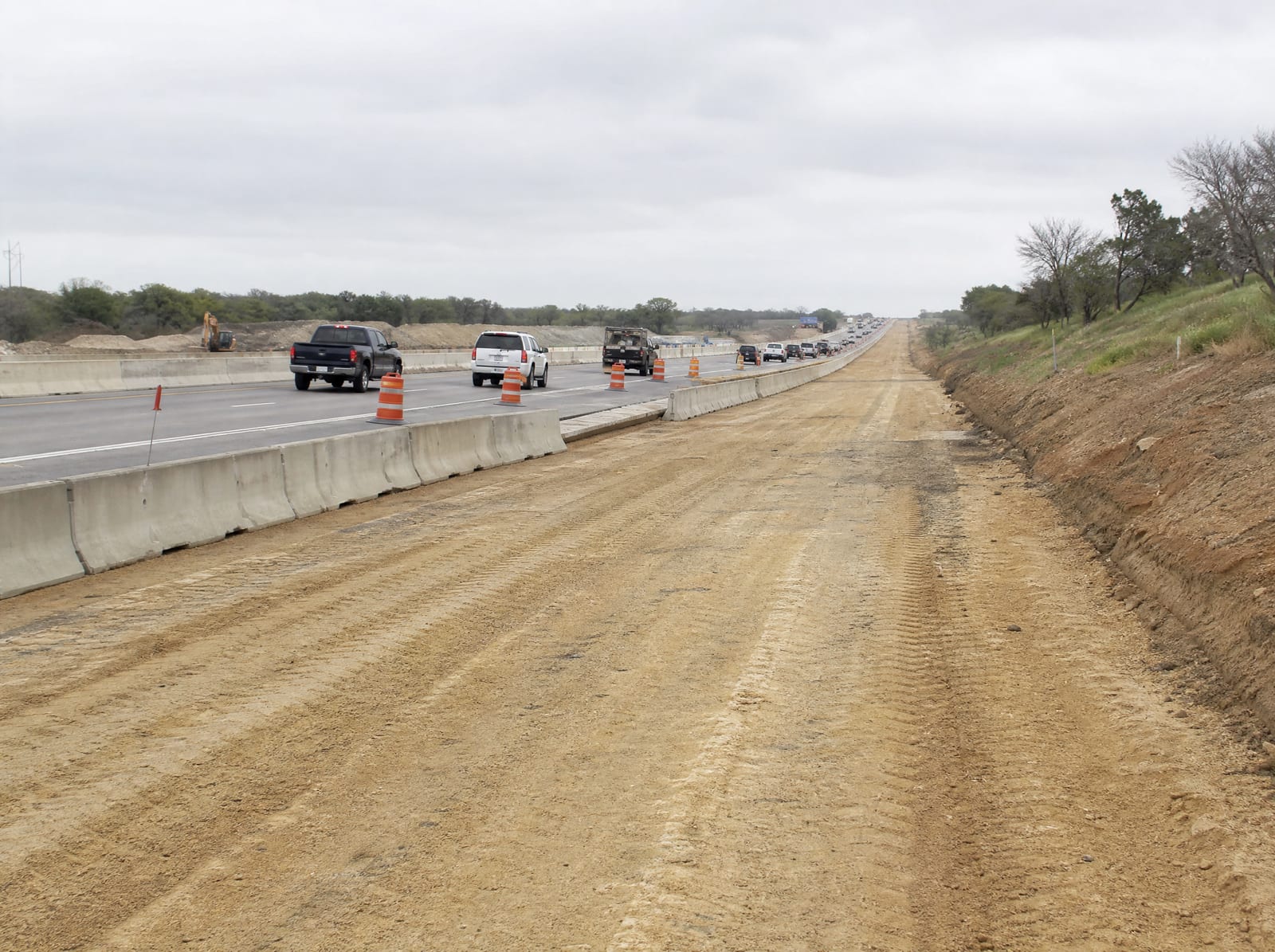 A Texas highway expansion in progress, a divided highway being widened with a graded shoulder, rows of orange barrels, and staged concrete barriers