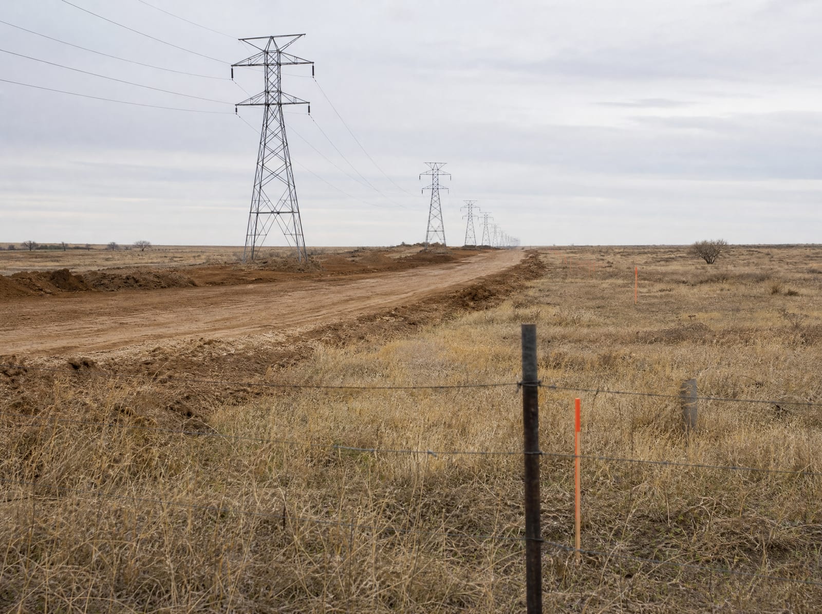 Rural Texas infrastructure development, a graded dirt access road and a line of electric transmission towers crossing dry ranchland behind a barbed-wire fence and survey stakes