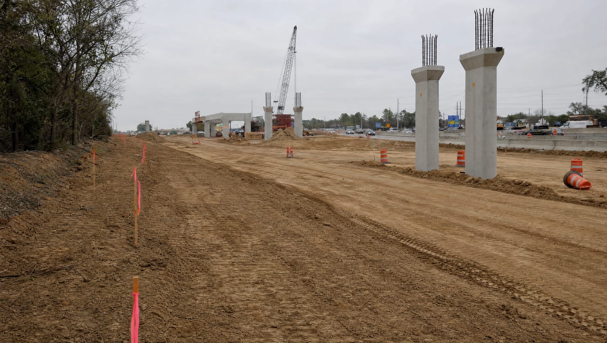 A major Texas highway-widening and interchange construction site with new concrete columns, graded earth, orange barrels, and survey stakes with right-of-way flags along the private-land edge