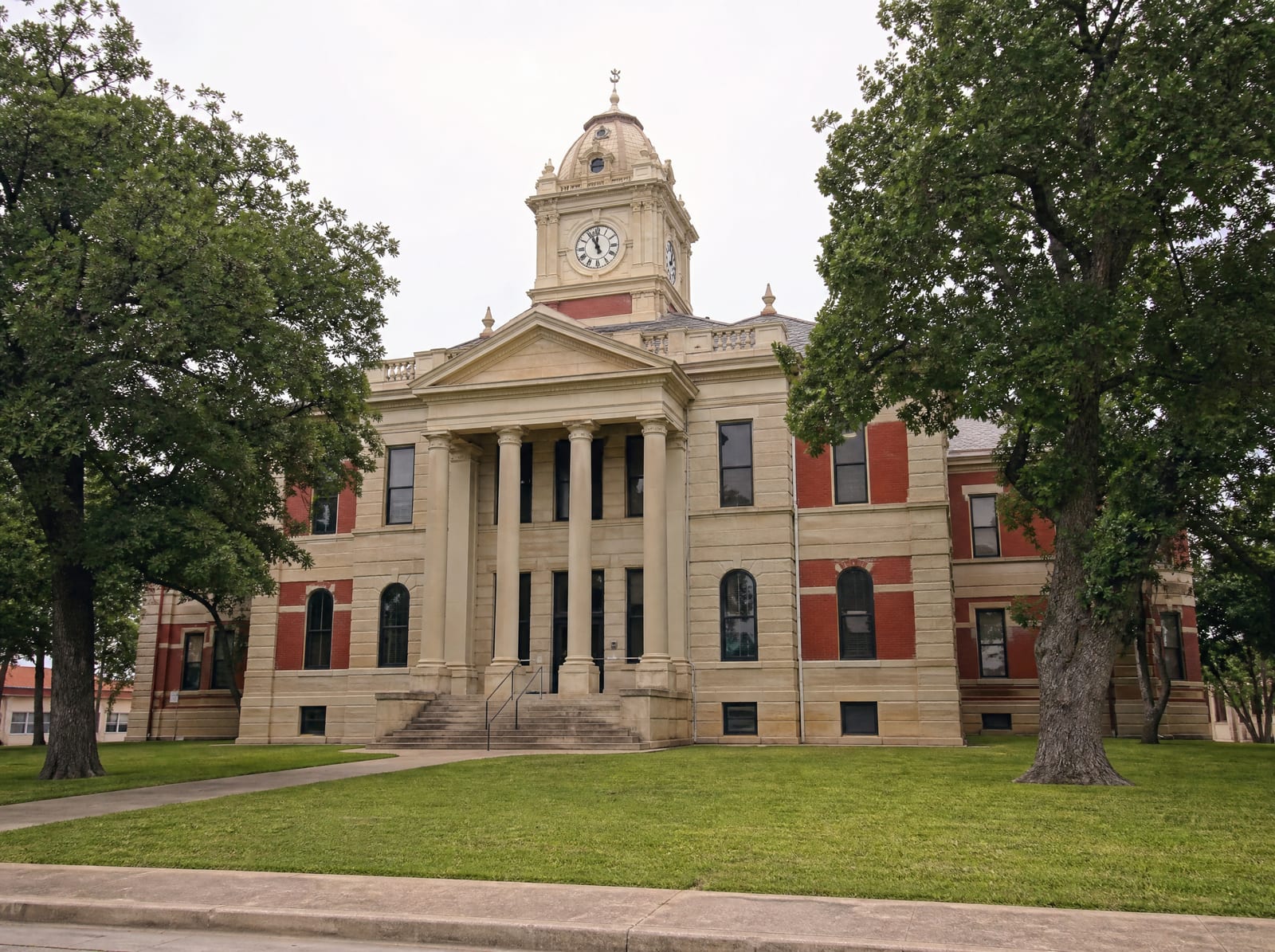A dignified historic Texas county courthouse of warm limestone and red brick with a clock tower, columns, and stone steps among mature live oaks, where guardianship and elder-law matters are heard