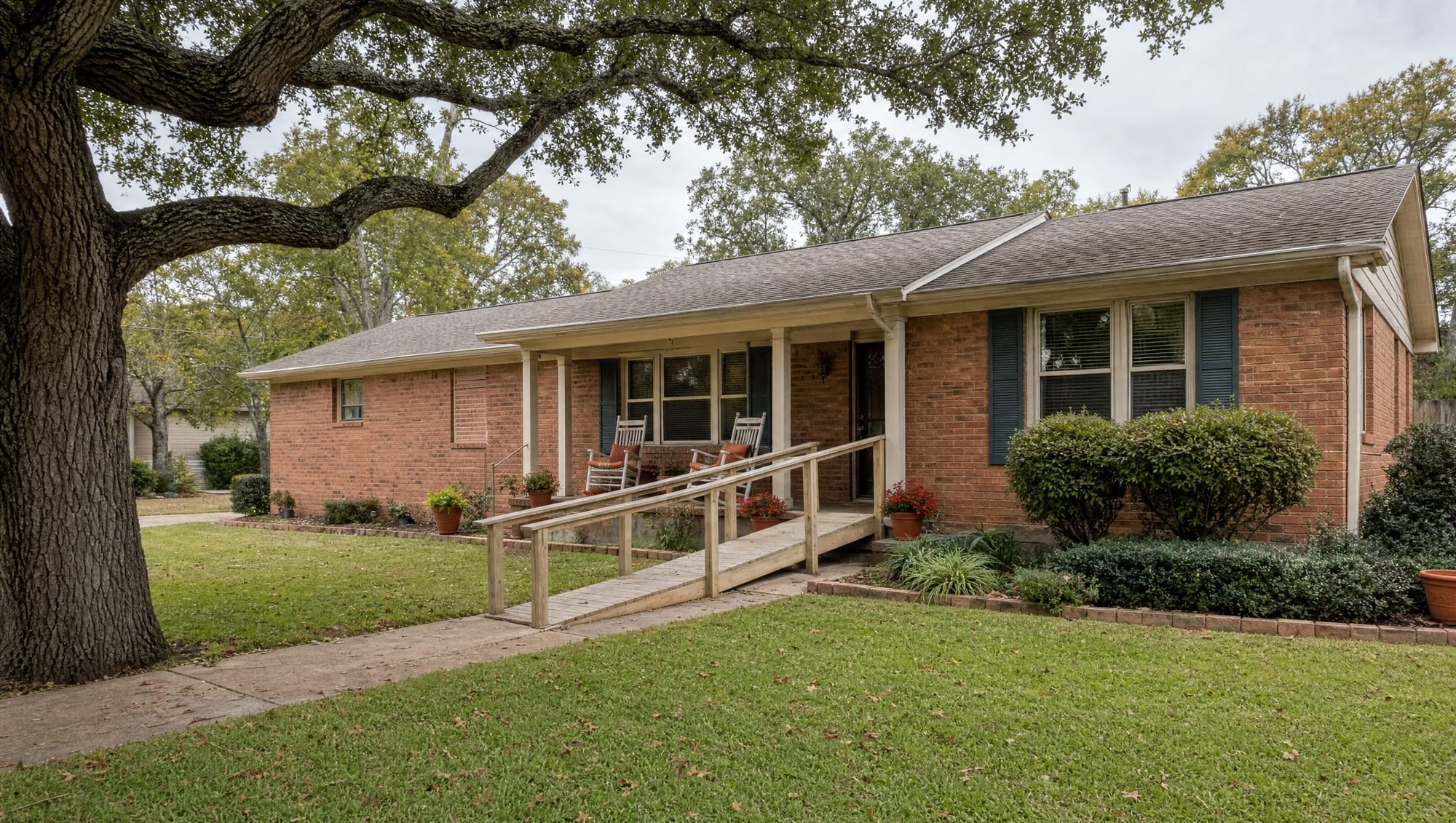 A welcoming established single-story red-brick Texas family home with a covered porch, two rocking chairs, potted flowers, and a gentle wheelchair-accessible wooden ramp to the front door, shaded by a mature live oak, a home where a longtime resident can age in place with dignity