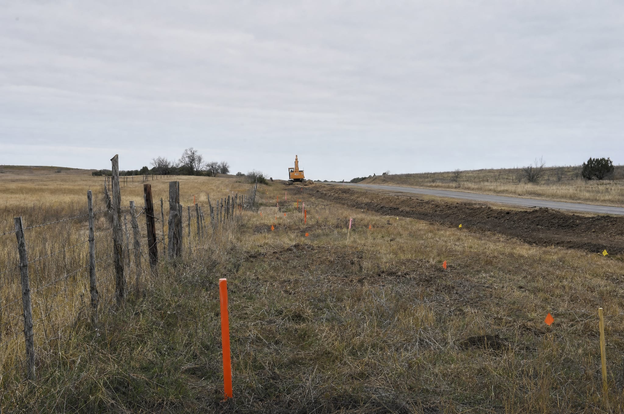 Orange survey stakes and marker flags in a field of dry grass beside a county road, with an excavator in the distance, as road construction encroaches on rural Texas land