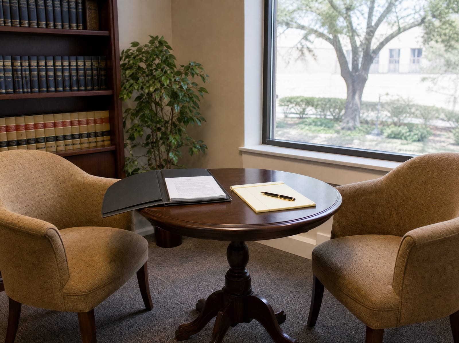 A warm law-office meeting corner with two upholstered chairs and a small table by a window, an open document folder and a legal pad, and a shelf of law books