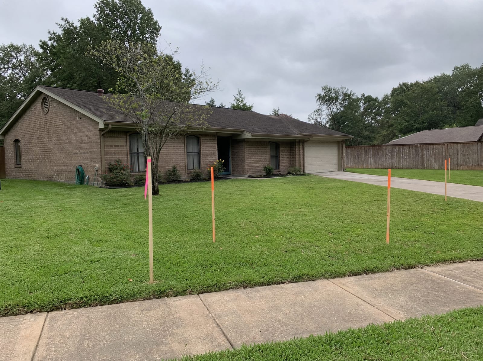 A modest single-story brick Texas home with a mown lawn and driveway, with orange survey stakes and a pink right-of-way flag in the front yard near the sidewalk