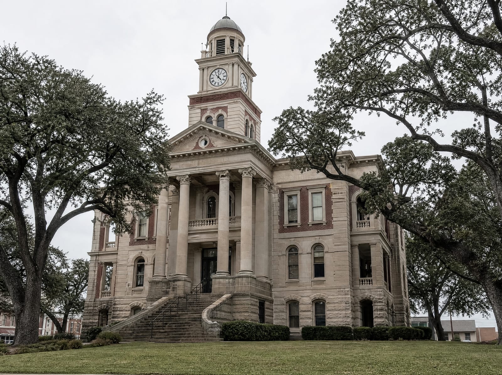 A historic limestone and brick Texas county courthouse with a clock tower and columns among live oaks