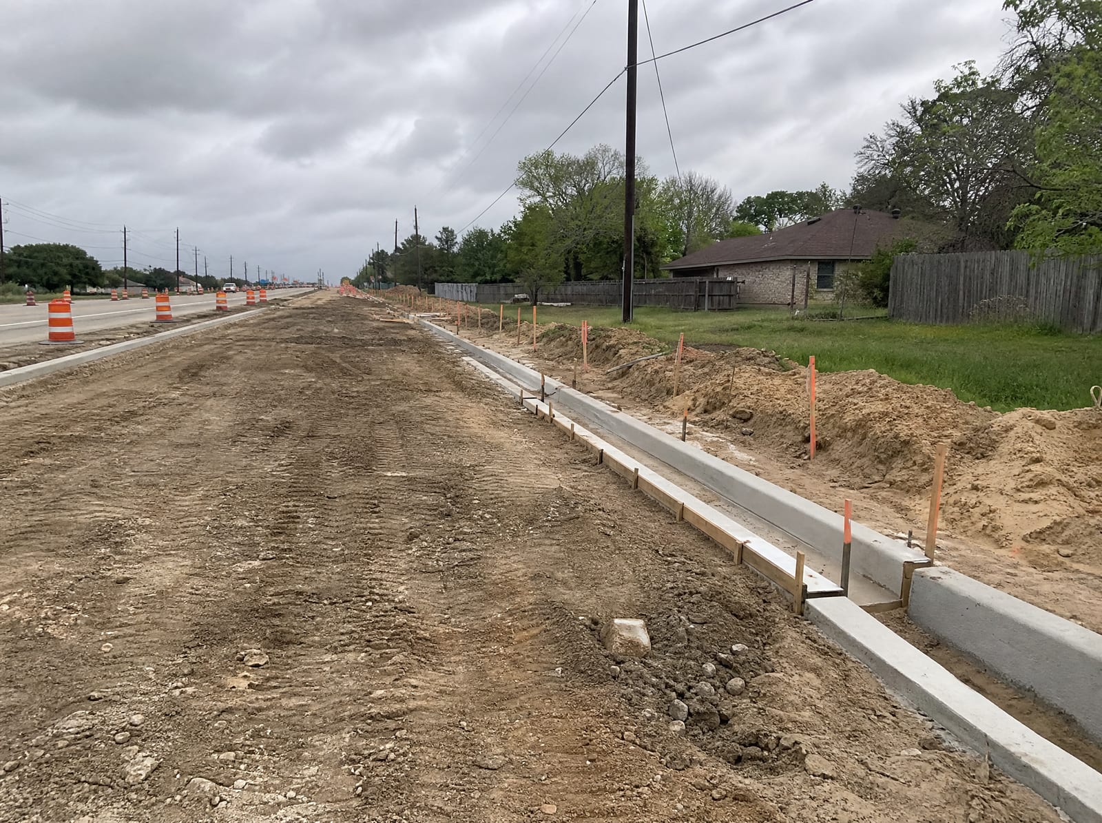 A road-widening construction zone beside Greater Houston property with orange barrels, graded dirt, and survey stakes along the property line