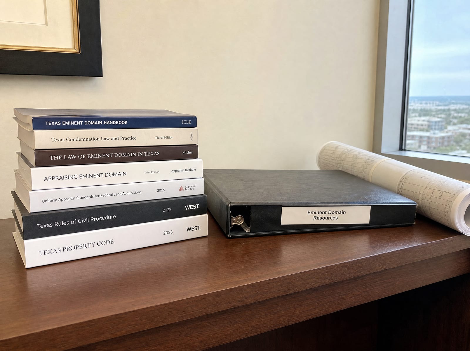 A neat stack of printed guides and a closed binder beside a rolled property map on a law-office desk, organized eminent-domain reference materials
