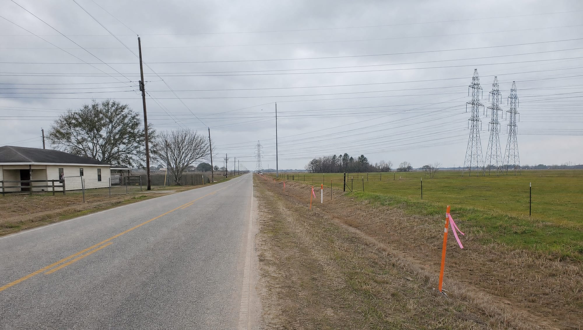 A two-lane rural Fort Bend County road with open pastureland and a fence on one side and a modest home on the other, survey stakes and right-of-way flags at the frontage, and transmission towers on the horizon