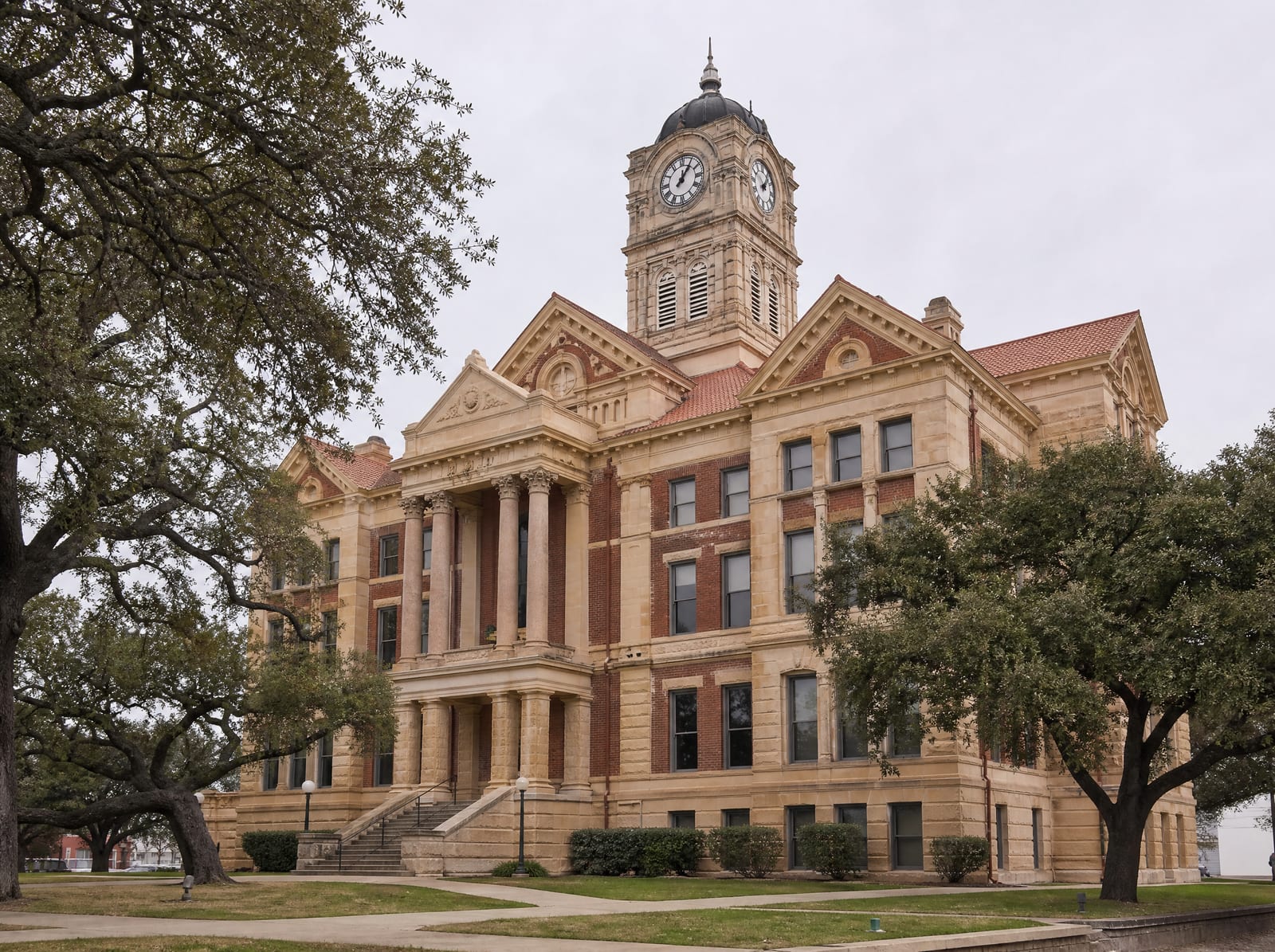 A dignified historic Texas county courthouse of warm limestone and red brick with a clock tower, columns, and stone steps among live oaks, where civil disputes are tried