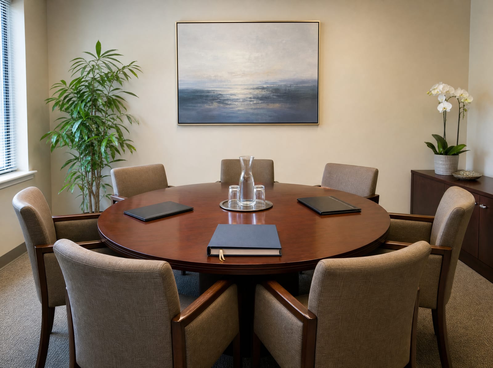 A neutral conference room with a round polished wood table, upholstered chairs, a settlement document, folders, and a water carafe, set for mediation or arbitration