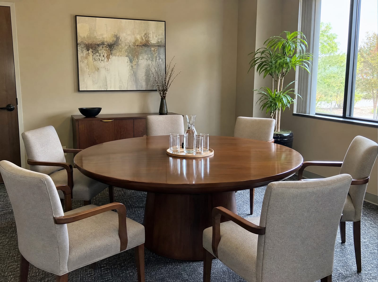 A neutral mediation conference room with a round polished wood table, comfortable upholstered chairs arranged evenly, a water carafe and glasses, and a plant by a window, set for a personal-injury mediation