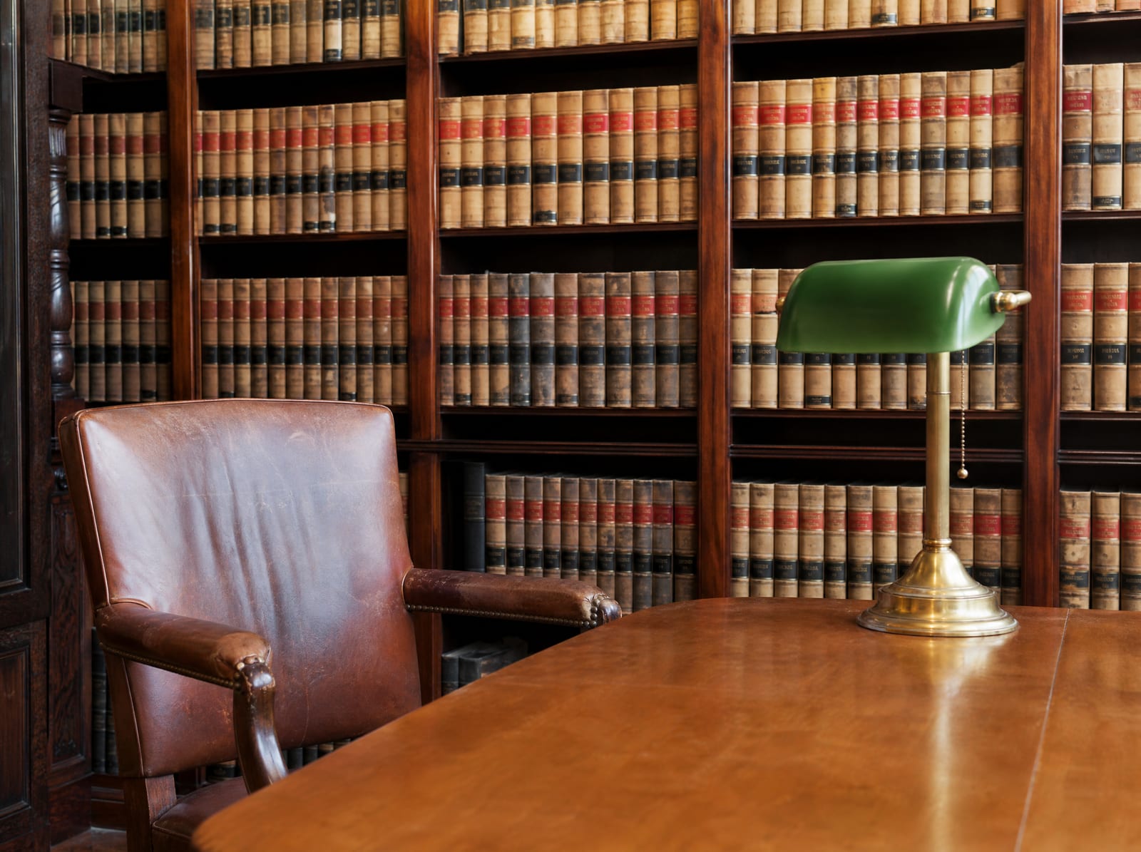 A distinguished law-firm library of well-worn bound law reporters with a wood reading table, a green-shaded brass banker's lamp, and a leather chair