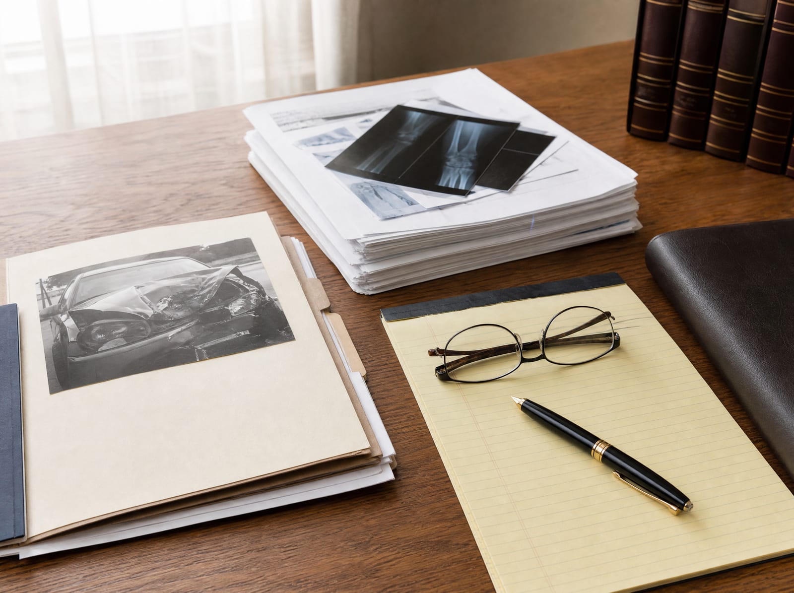 A personal-injury case file and folder, a set of medical records, a legal pad with a fountain pen, and reading glasses on a warm wood desk, as an attorney builds a client's injury claim