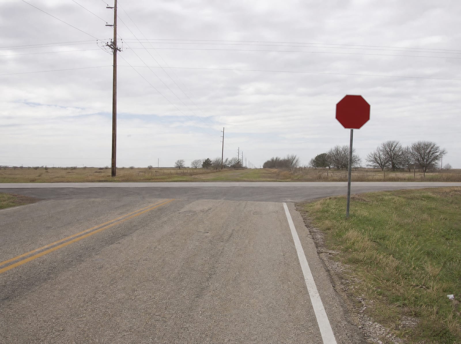 A quiet two-lane Texas road at a rural intersection with a stop sign and utility poles, empty and calm under a soft sky, the kind of setting where a serious accident can happen