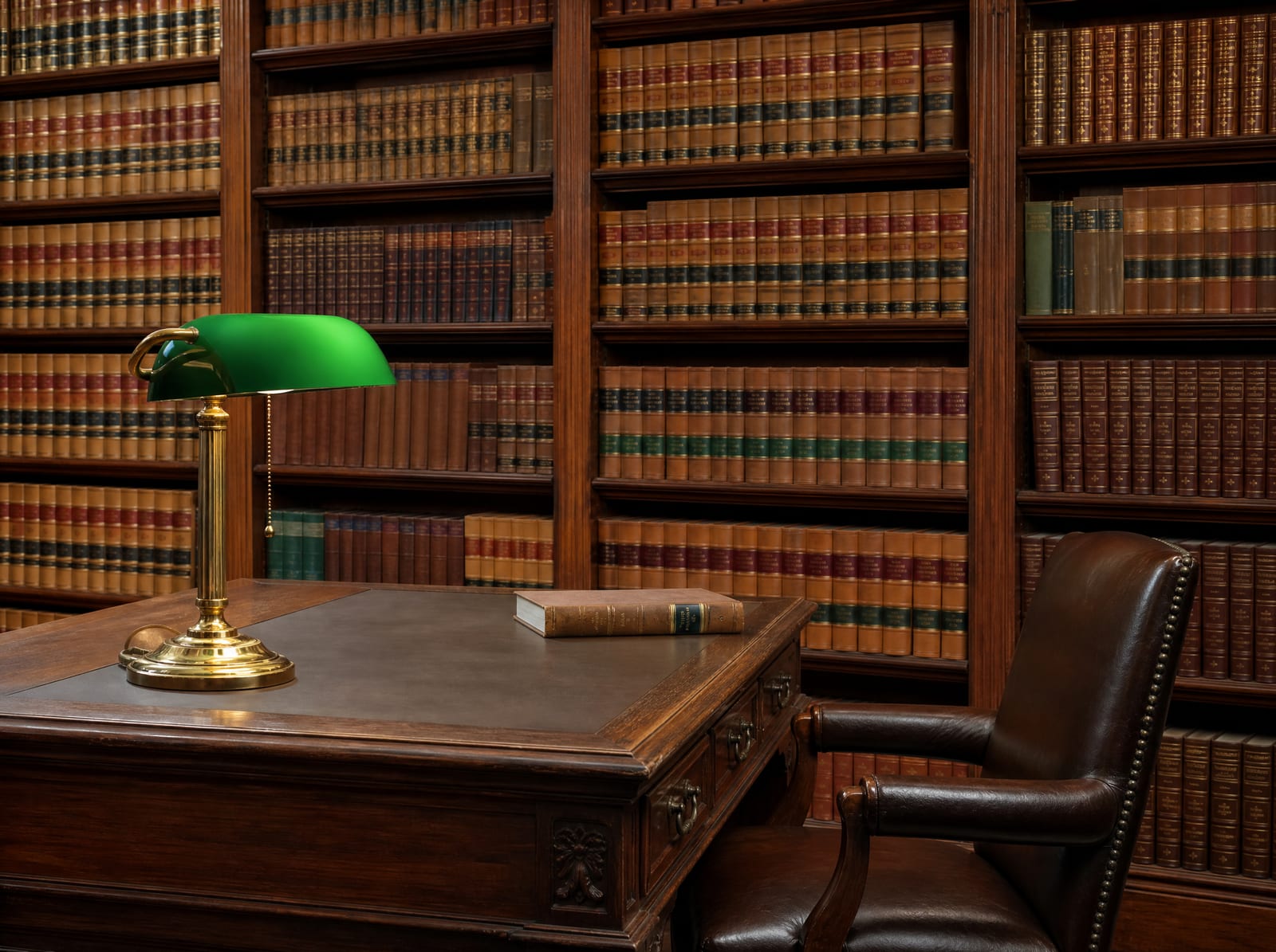 A distinguished law-firm library of well-worn bound law reporters with a wood reading table, a green-shaded brass banker's lamp, and a leather chair, reflecting long experience