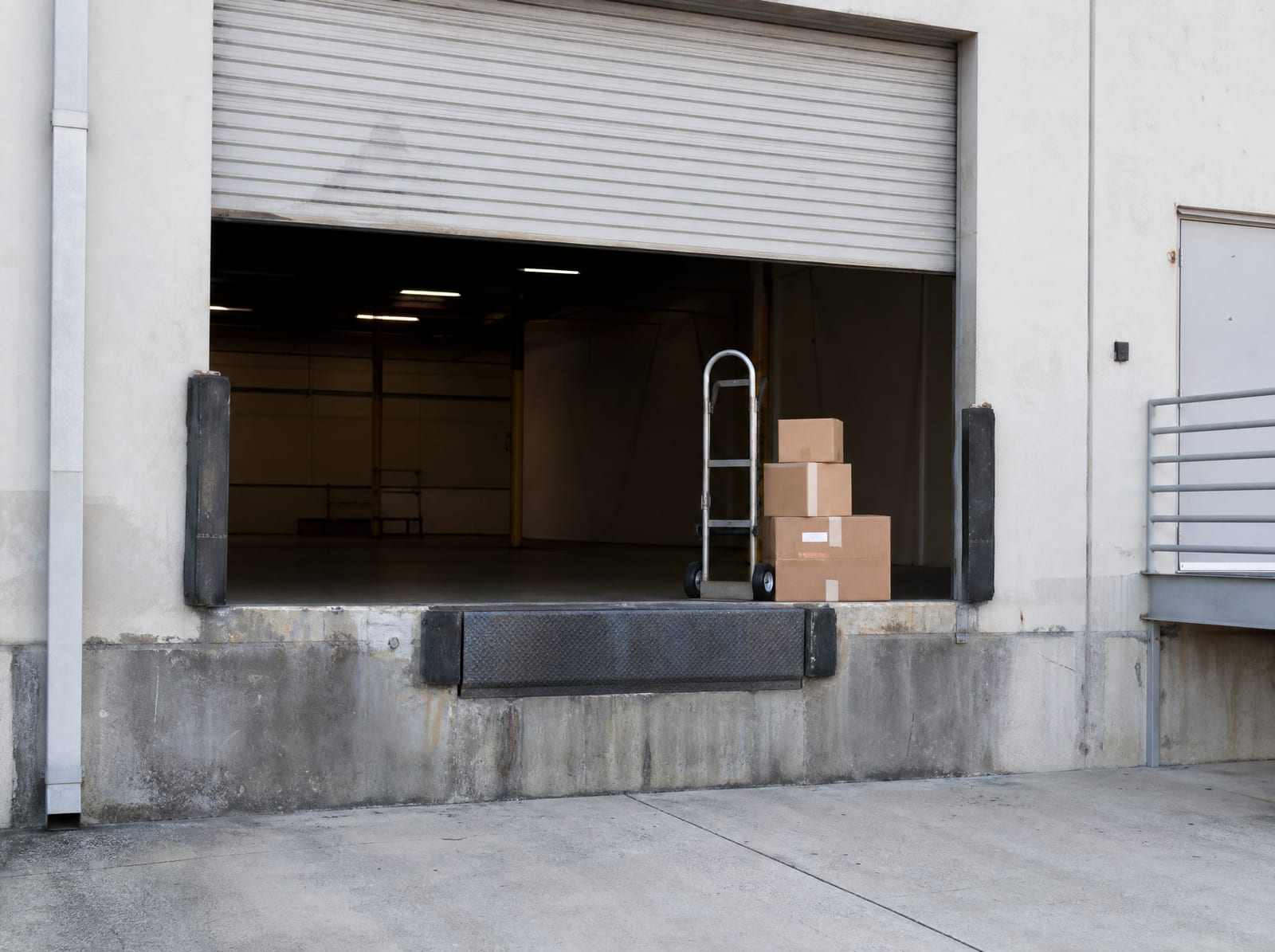 A commercial loading dock with a roll-up door, stacked moving boxes, and a hand truck, suggesting a business relocating after a taking