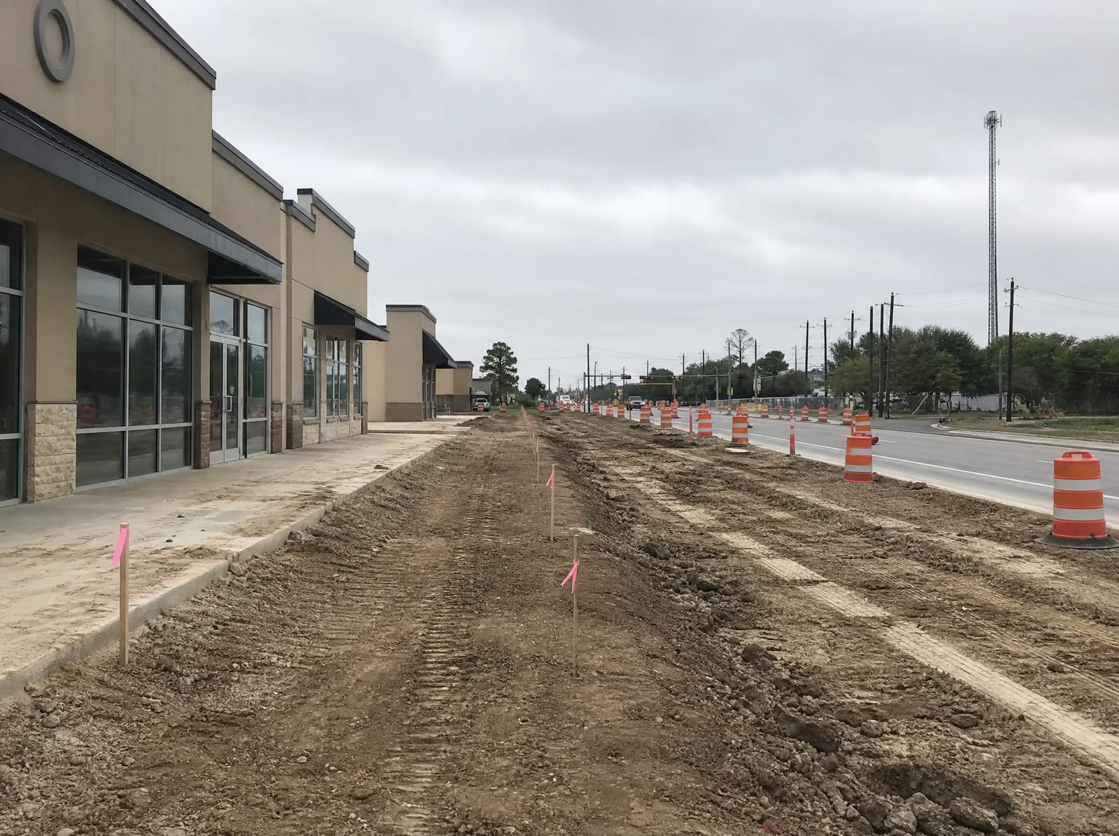 A Texas commercial strip center beside a road-widening construction zone with cones, graded dirt, and survey stakes along the property line