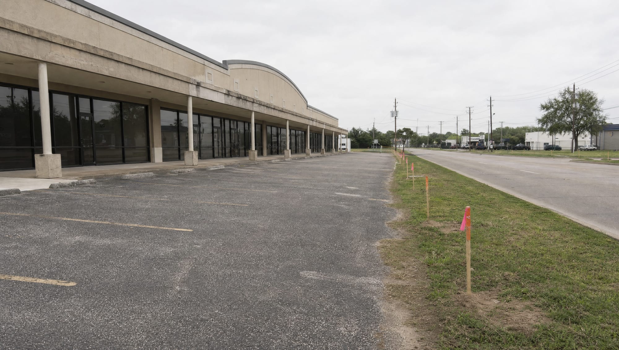 A Texas multi-tenant retail strip center with a covered walkway, glass storefronts, and a parking lot along a frontage road, with orange survey stakes and pink right-of-way flags along the street edge under an overcast sky