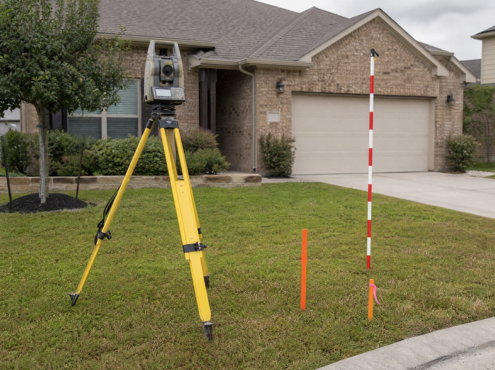 A surveyor's tripod and total station set up on the front lawn of a suburban brick home, with survey stakes and a leveling rod, during a property analysis