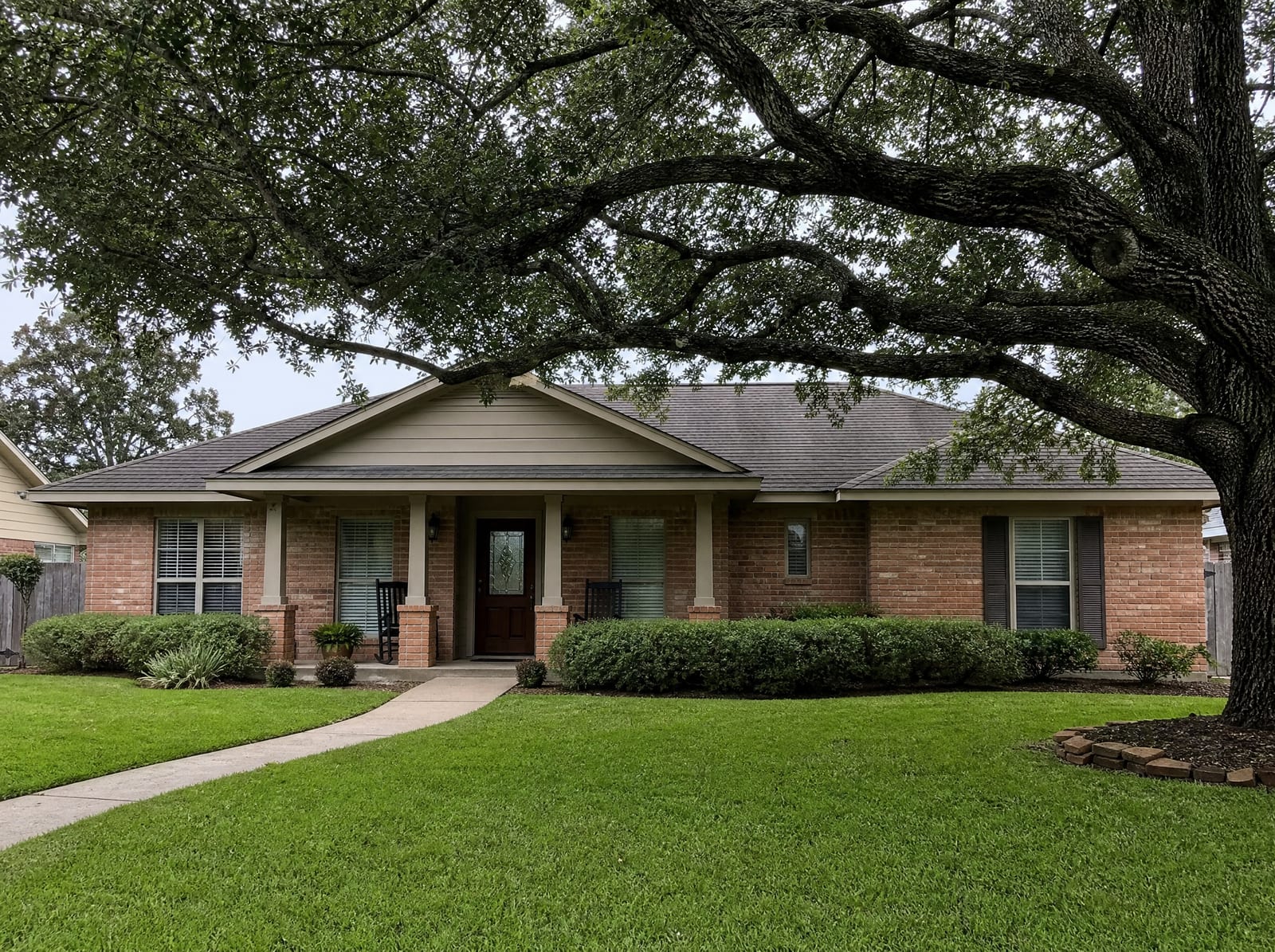 A well-kept single-story brick Texas home with a covered porch and manicured lawn shaded by a mature live oak in a quiet suburban neighborhood