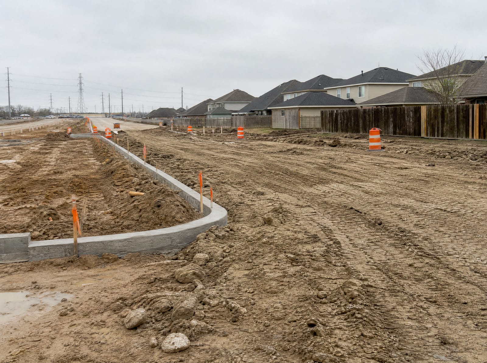 A road-widening construction zone with graded dirt, orange cones, and survey stakes beside a Greater Houston residential neighborhood