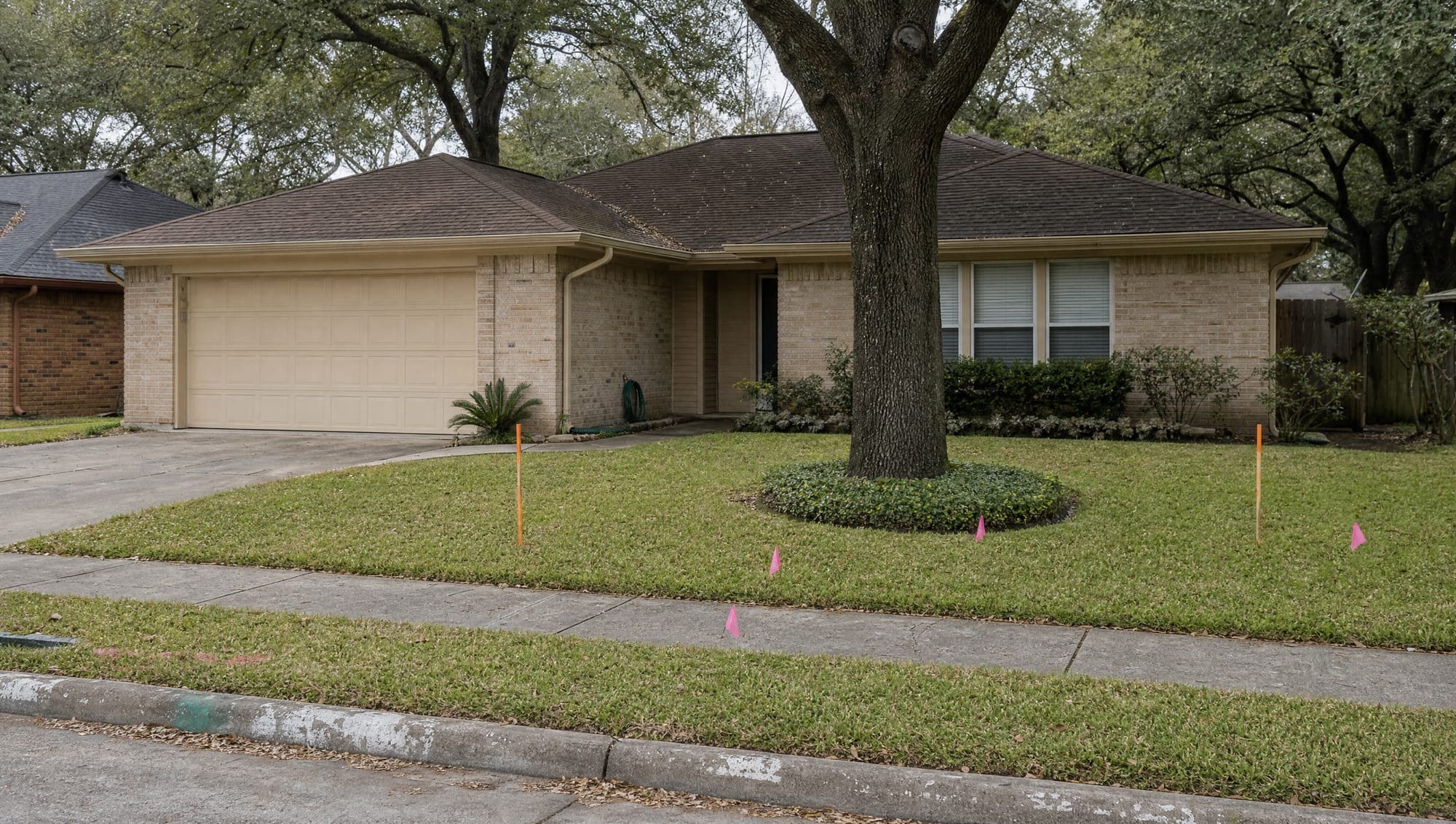 A single-story brick Texas family home in an established Greater Houston neighborhood, with orange survey stakes and pink right-of-way flags in the front yard and along the sidewalk under an overcast sky