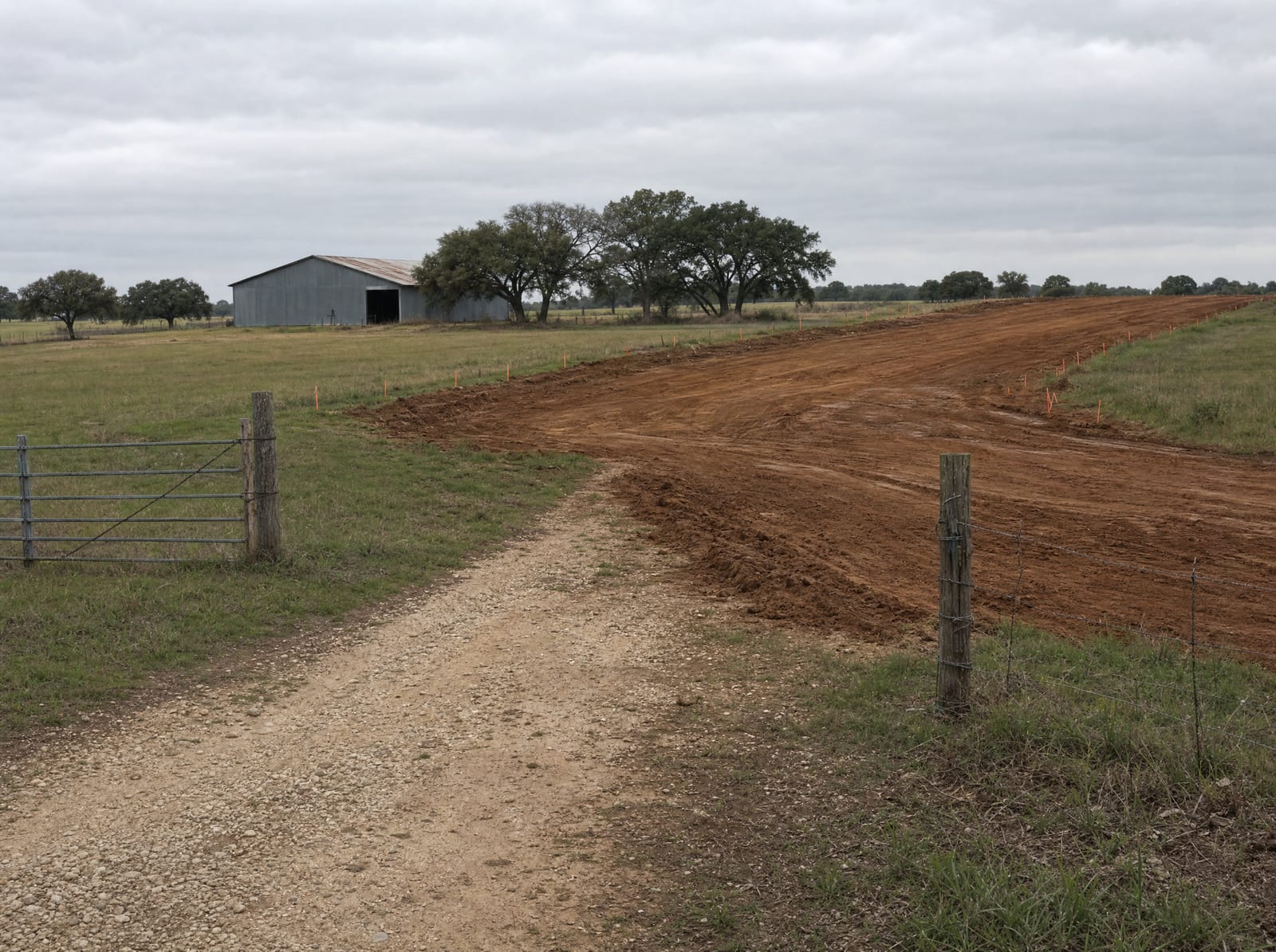 A rural Texas property where a cedar-post fence line and gravel drive are interrupted by a freshly graded dirt right-of-way cut, with survey markers, a barn, and live oaks beyond