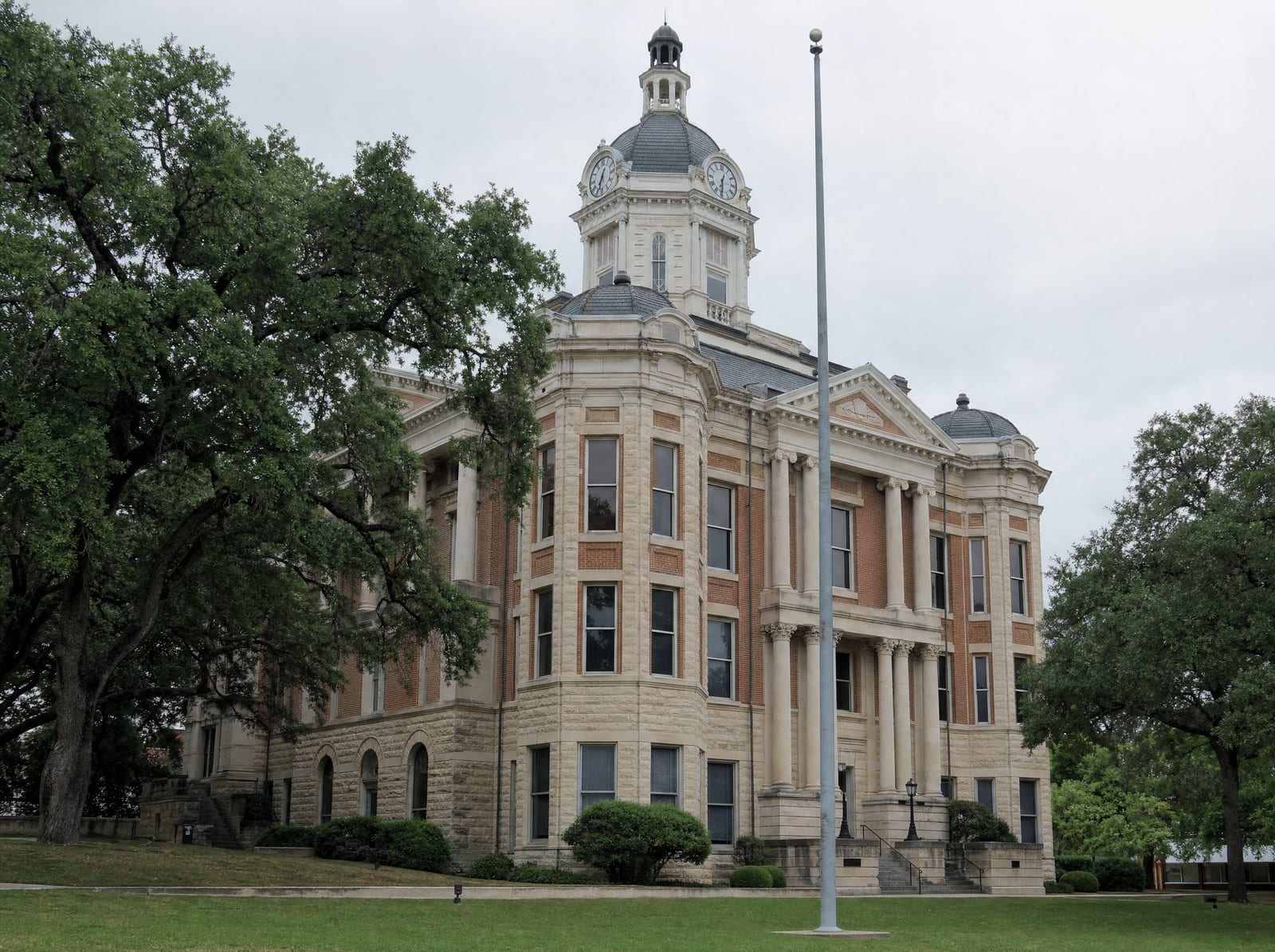 A historic limestone and brick Texas county courthouse with a clock tower and columns among mature live oaks, where Special Commissioners hearings are held