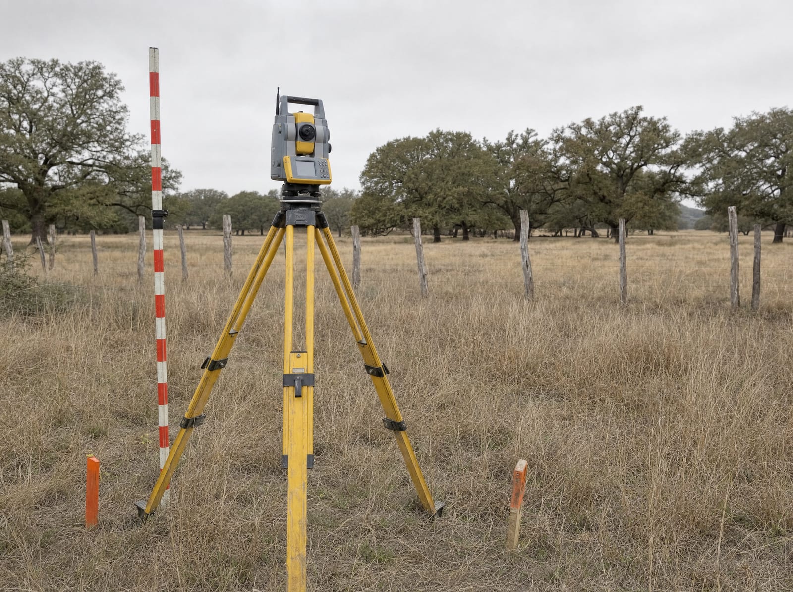 A surveyor's tripod and total station set up in an open Texas field with survey stakes and a leveling rod, a cedar-post fence and live oaks behind, for an independent land appraisal