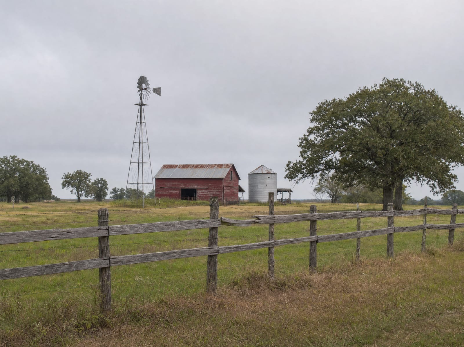 Open Fort Bend County ranchland with scattered live oaks, a weathered red barn and grain silo, and an old windmill behind a cedar-post fence, the landowner's livelihood