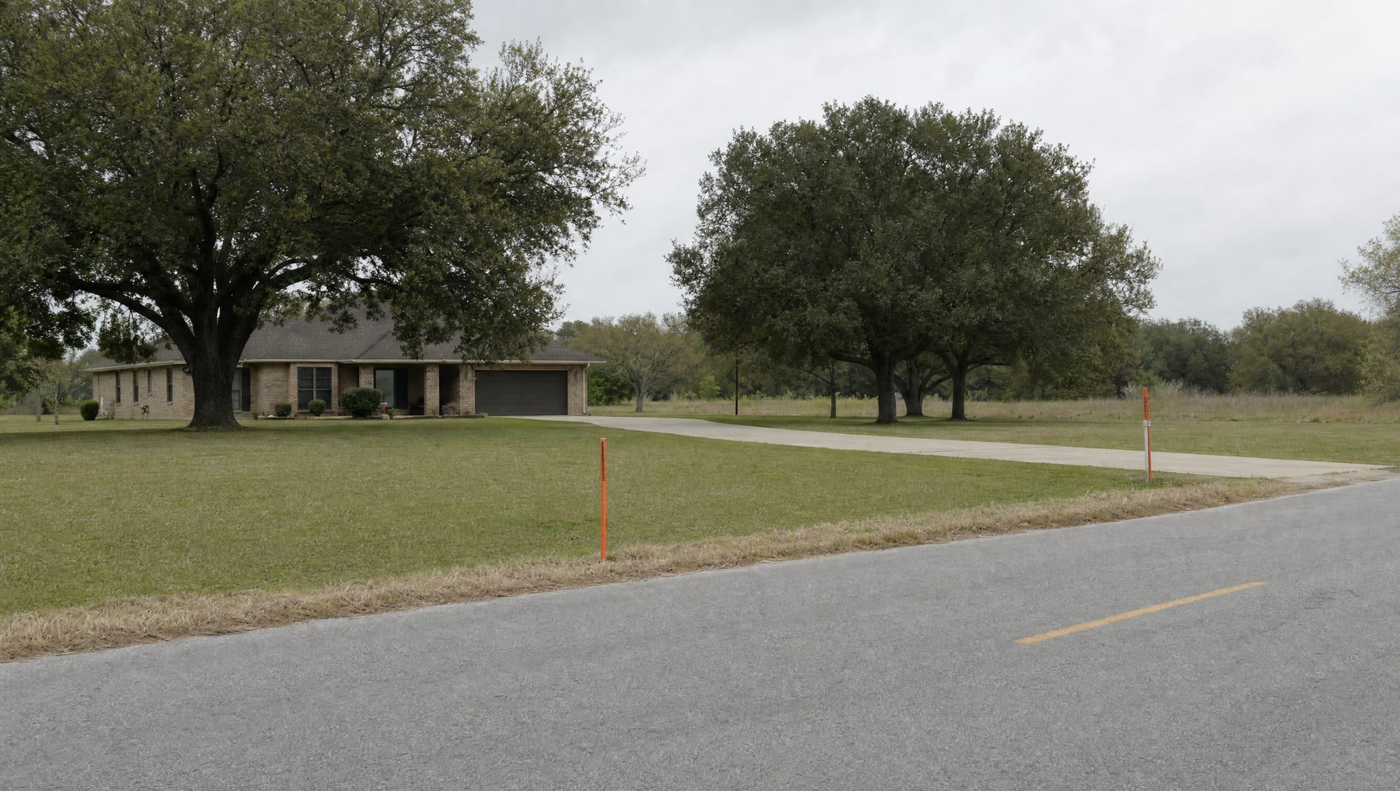 A modest single-story Texas home on a generous lot with a mown lawn, mature live oaks, and open land along a quiet county road, with survey stakes at the road frontage, the property whose just compensation is at stake