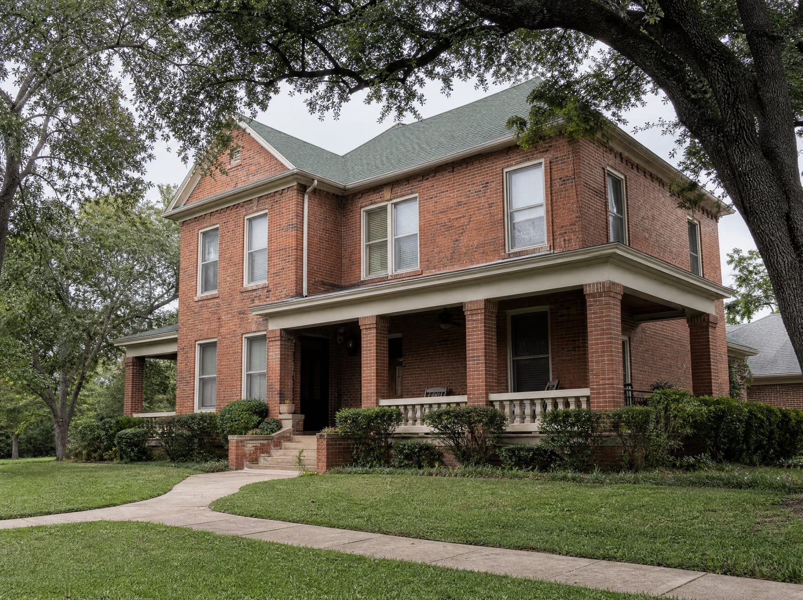 A warm, established two-story brick Texas family home with a covered porch and mature shade trees, the family estate at the heart of an inheritance dispute