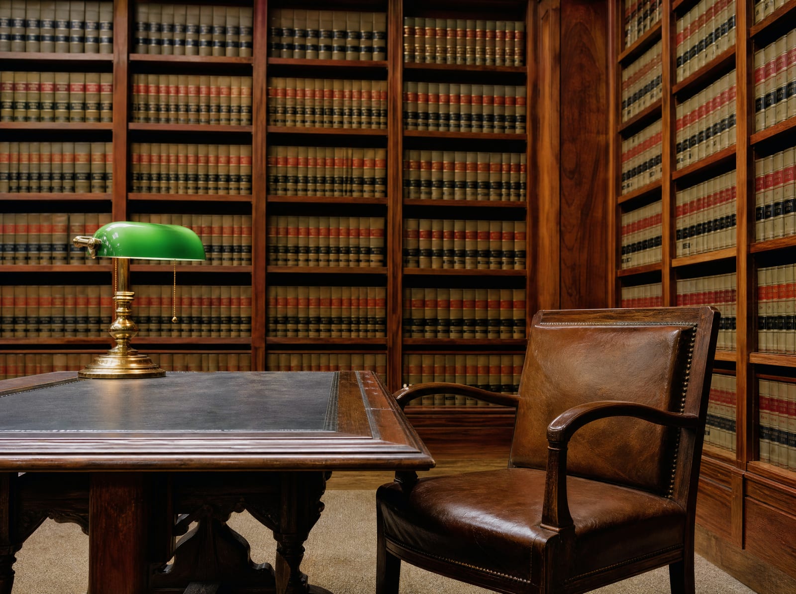A dignified law-firm library with floor-to-ceiling shelves of bound law reporters, a wood reading table with a green-shaded brass banker's lamp, and a leather chair