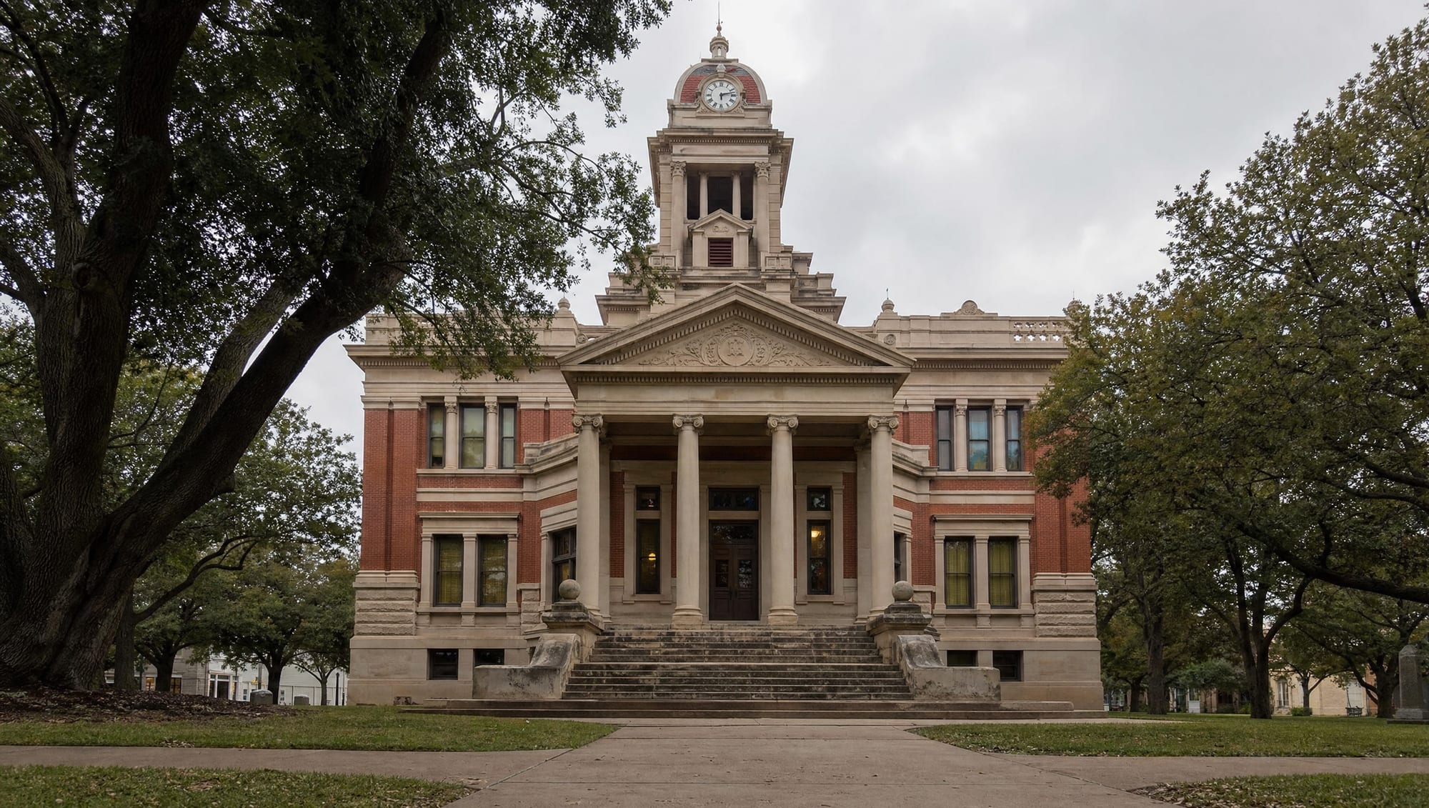 A dignified historic Texas county courthouse of warm limestone and red brick with a clock tower, columns, and broad stone steps among live oaks, where will contests and estate disputes are decided